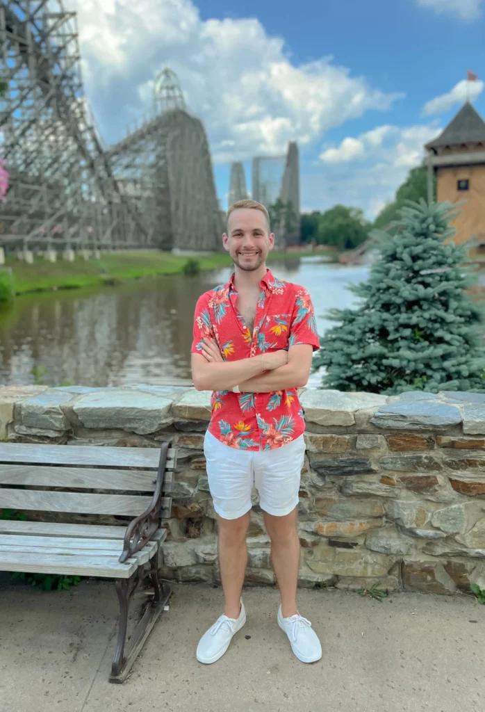 Light skinned man smiling and standing in front of the lake at Six Flags Great Adventure. El Toro, Kingda Ka, and Runaway Mine Train roller coasters are in the distance