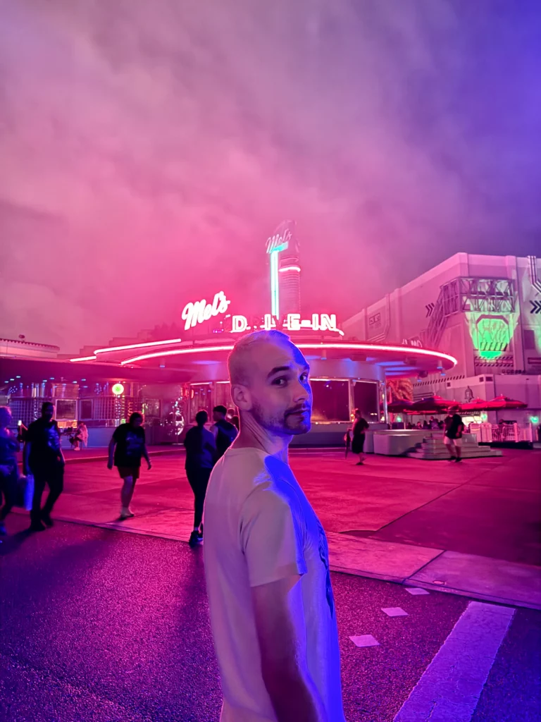 Light skinned man looking over shoulder with neon lights of Mel's Die In behind him lighting him up at night