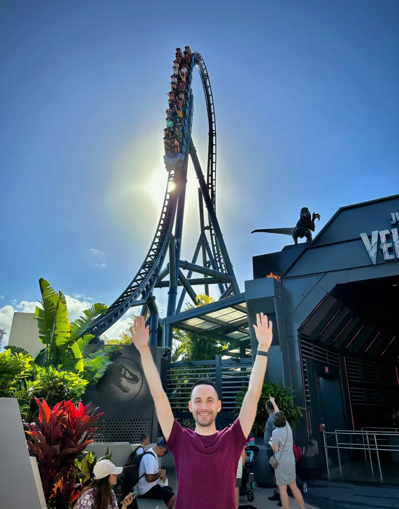 Light skinned man standing in front of VelcicCoaster entrance at Universal Islands of Adventure while a train goes down the top hat behind him